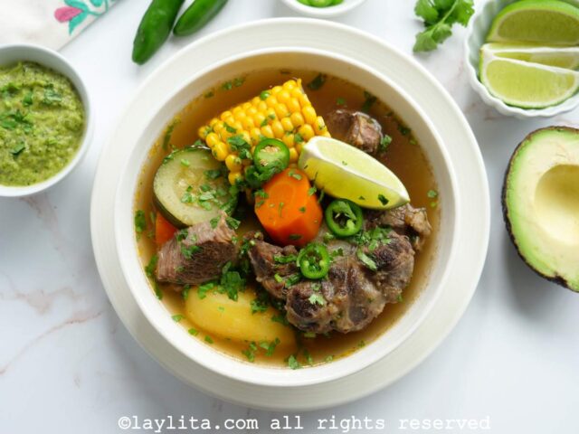 Overhead shot of caldo de res in a white bowl with lime, avocado and jalapeño salsa on the side