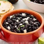 A reddish bowl filled with black beans garnished with crumbled cheese and cilantro, with tortillas and avocados on the side