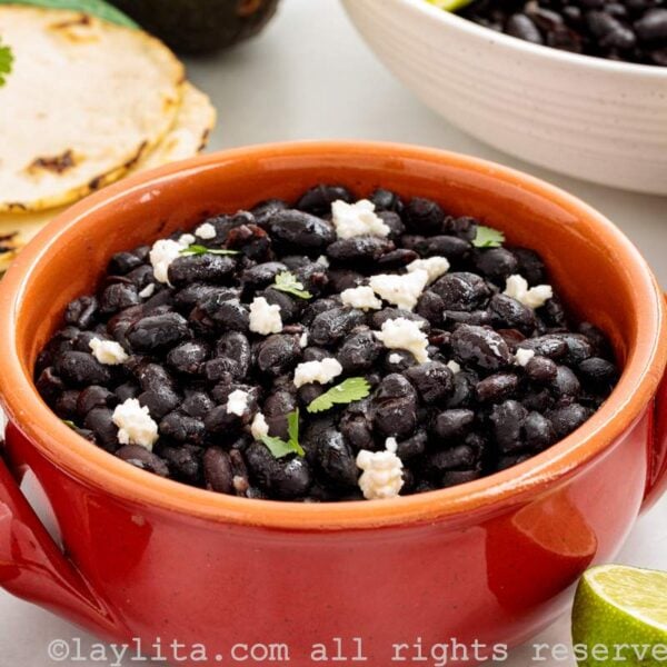 A reddish bowl filled with black beans garnished with crumbled cheese and cilantro, with tortillas and avocados on the side