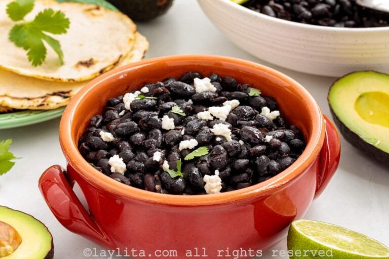 A reddish bowl filled with black beans garnished with crumbled cheese and cilantro, with tortillas and avocados on the side
