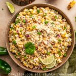 Mexican corn salad in a brown bowl on a countertop, next to the bowl are jalapeños, cilantro leaves, a small bowl with crumbled cheese