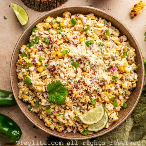 Mexican corn salad in a brown bowl on a countertop, next to the bowl are jalapeños, cilantro leaves, a small bowl with crumbled cheese