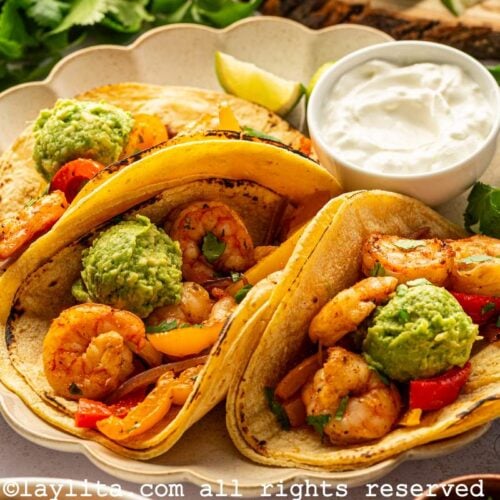Side view of three Shrimp Fajita tacos topped with mashed avocado on a plate with a bowl of sour cream, on the side there are avocado halves and cilantro sprigs