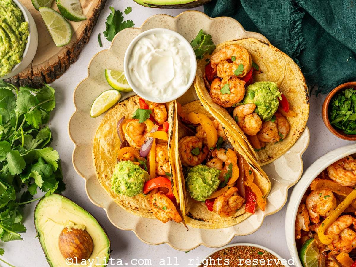 View of three shrimp tacos with veggies, and mashed avocado on top, with a bowl of sour cream next to them