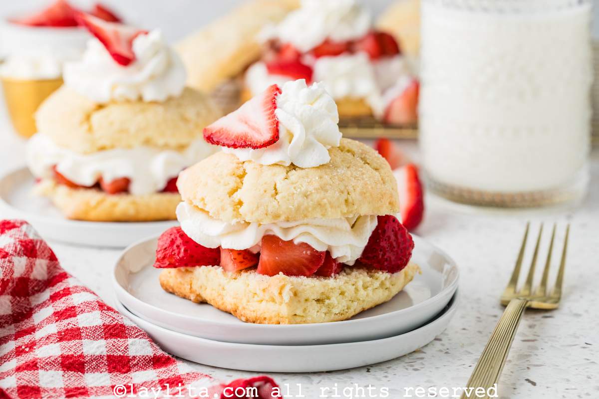 A strawberry shortcake on a plate with several more in the background, next to a red and white kitchen napkin and a fork