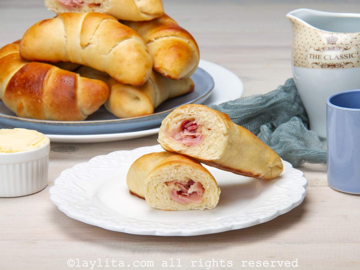 One ham cachito broken in half to show the filling on a white plate, with more bread rolls in the background