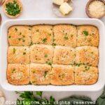A dozen cheese stuffed dinner rolls in a white baking dish, with chopped herbs and butter on the side