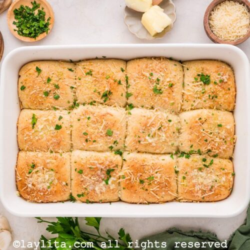 A dozen cheese stuffed dinner rolls in a white baking dish, with chopped herbs and butter on the side