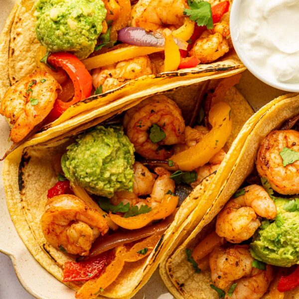 Close-up view of three tacos filled with shrimp, veggies, and mashed avocado with a bowl of sour cream next to them
