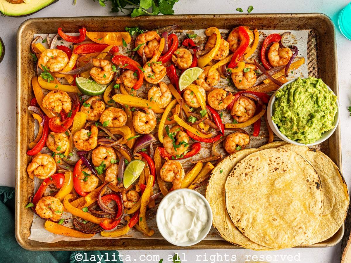 Overhead view of a baking sheet pan filled with roasted shrimp and sliced veggies, with corn tortillas, mashed avocado and sour cream on the side of the pan