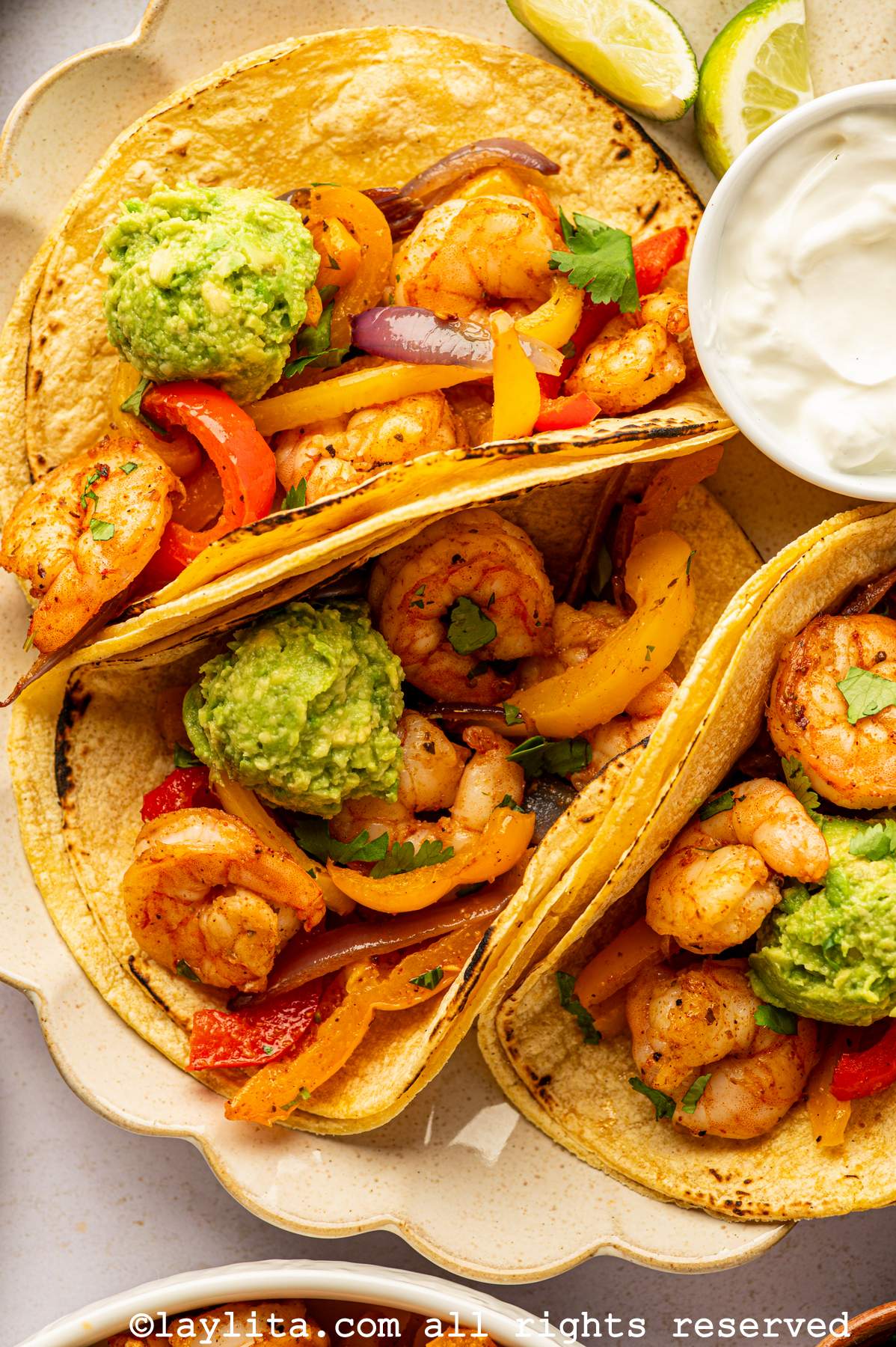 Close-up view of three tacos filled with shrimp, veggies, and mashed avocado with a bowl of sour cream next to them