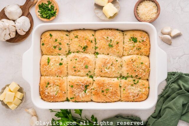 Dinner rolls with parmesan and parsley on top on a counter next to bowls of parmesan and chopped parsley