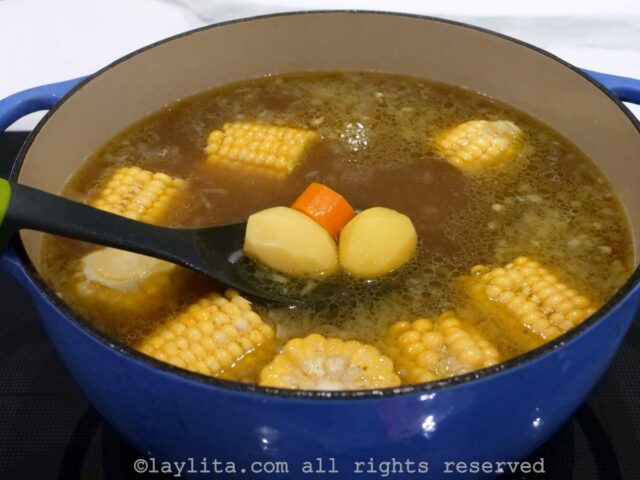 A spoon showing potatoes and carrots cooking with corn in a soup pot