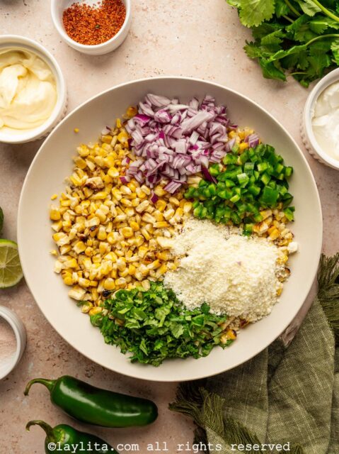 A white salad bowl with grilled corn kernels, diced onion, diced jalapeño, chopped cilantro and crumbled cheese