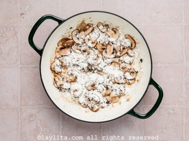 Flour being sprinkled over cooked mushrooms in a skillet