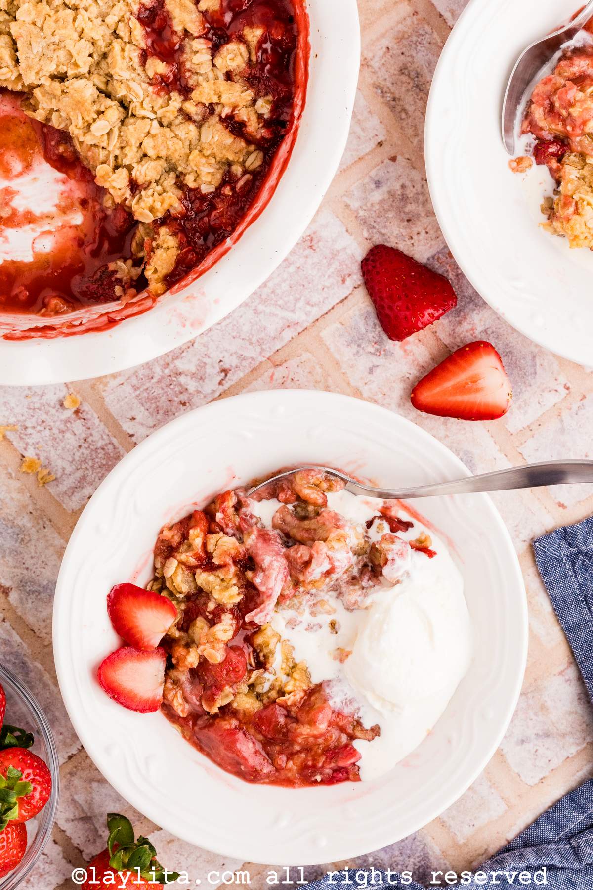Overhead shot of a portion of strawberry crisp in a white bowl with a scoop of vanilla ice cream, with more strawberry crisp in the background