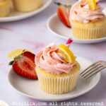 A strawberry lemonade cupcake decorated strawberry frosting on a white plate, with a strawberry half and a fork next to it