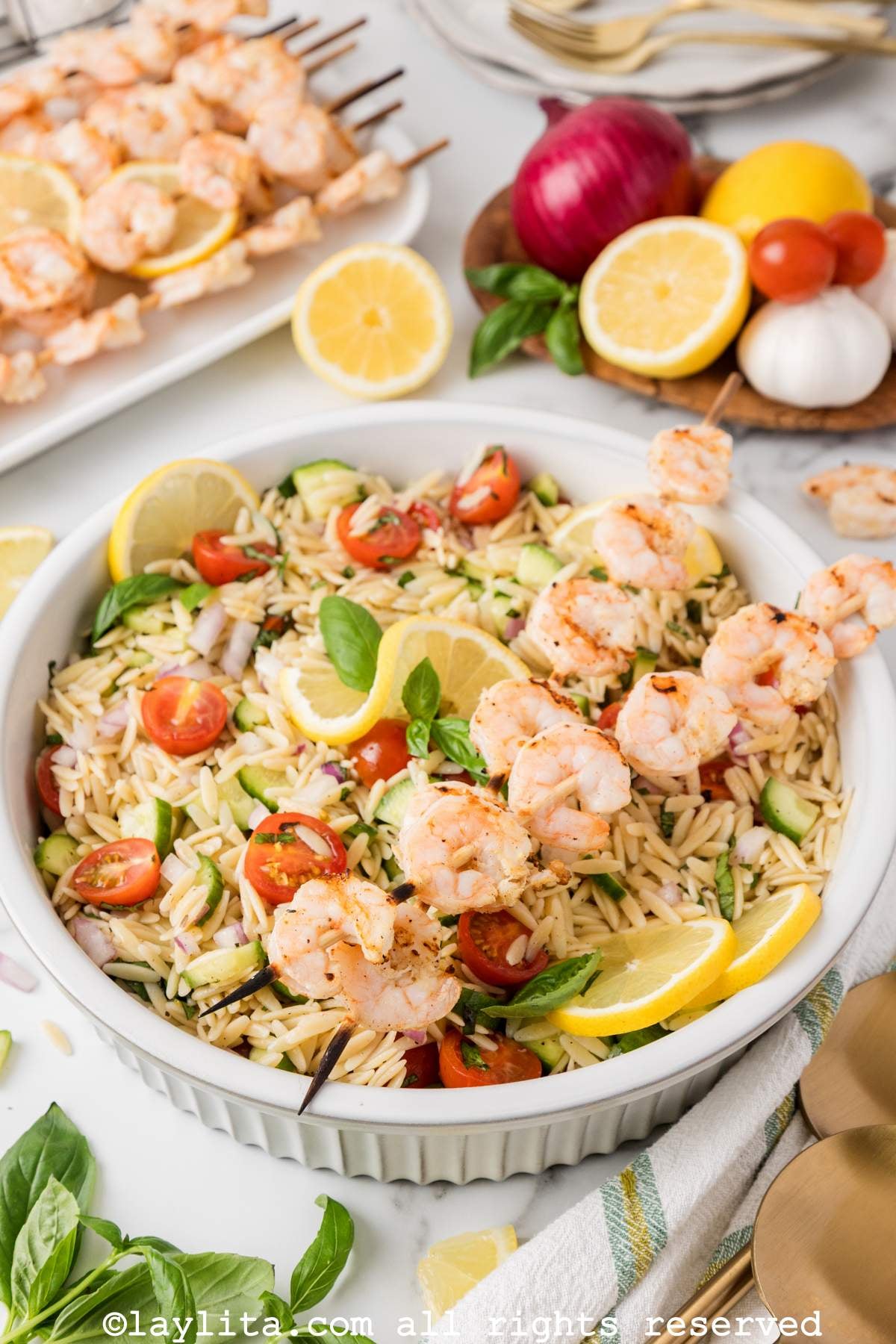 Side view of a bowl of orzo pasta salad topped with grilled shrimp, with more shrimp and vegetables in the background
