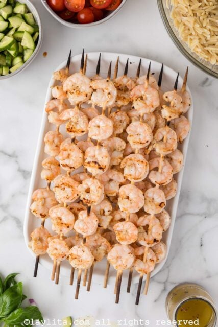 A platter of lemon marinated grilled shrimp skewers on a kitchen counter next to ingredients for orzo pasta salad