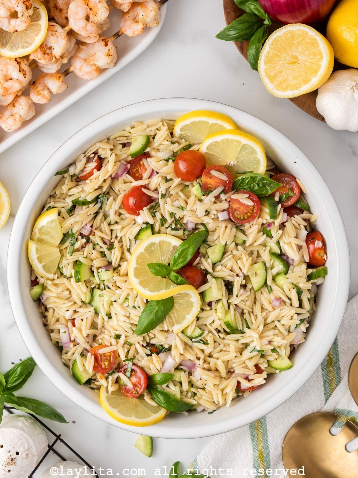 Overhead view of a large white bowl full of orzo pasta salad garnished with lime slices and basil leaves, on the side there is a partial view of a plate of grilled shrimp skewers