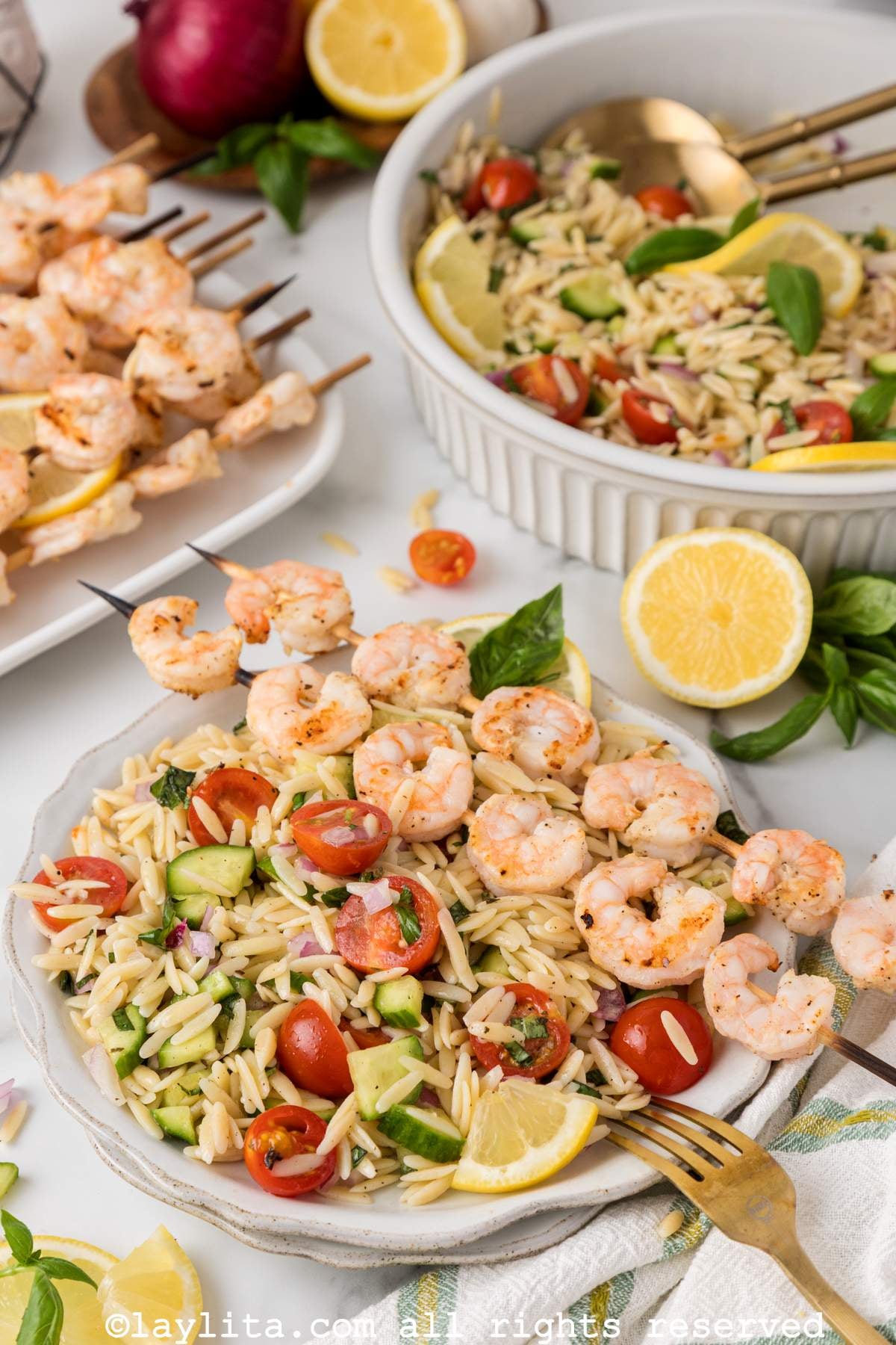 A plate of lemon orzo pasta salad with two grilled shrimp skewers on top, with more shrimp and the bowl of salad in the background