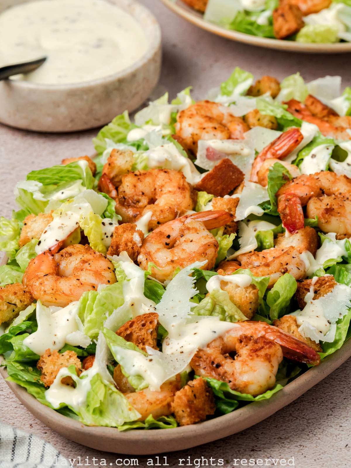 View of a platter full of Caesar Salad topped with shrimp, next to a bowl of homemade dressing