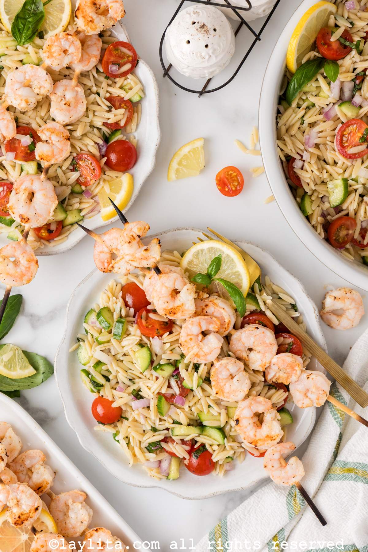 Overhead view of two white plates with orzo pasta salad and grilled shrimp skewers on top, with more salad and shrimp skewers on the sides