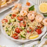Lemon orzo pasta salad with vegetables and grilled shrimp on a white plate with more grilled shrimp in the backgroun