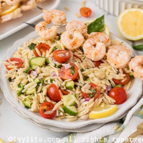 Lemon orzo pasta salad with vegetables and grilled shrimp on a white plate with more grilled shrimp in the backgroun