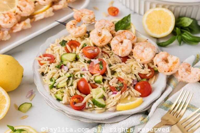Lemon orzo pasta salad with vegetables and grilled shrimp on a white plate with more grilled shrimp in the backgroun