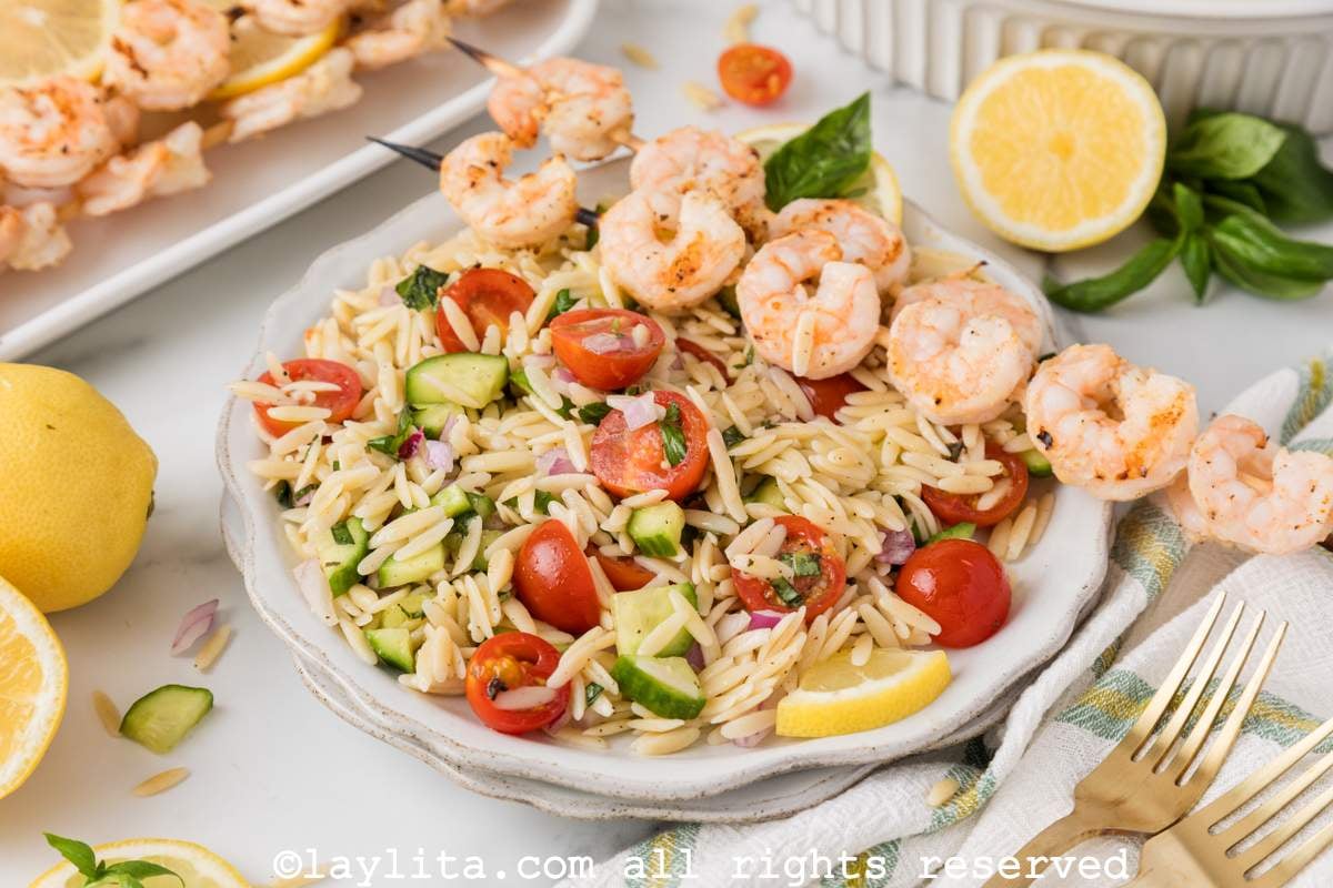 Lemon orzo pasta salad with vegetables and grilled shrimp on a white plate with more grilled shrimp in the backgroun