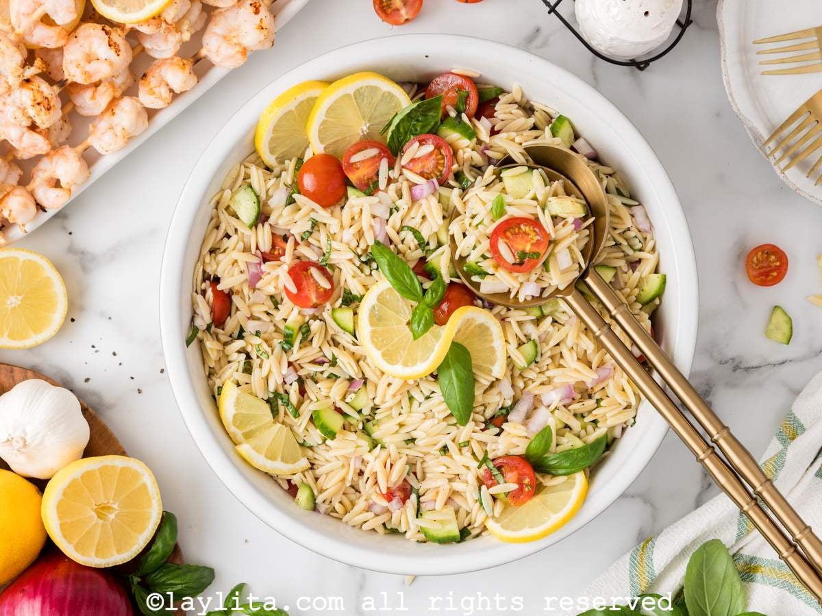 Overhead view of a bowl of orzo salad with tomato and cucumber, there is a large serving spoon in the middle of the salad