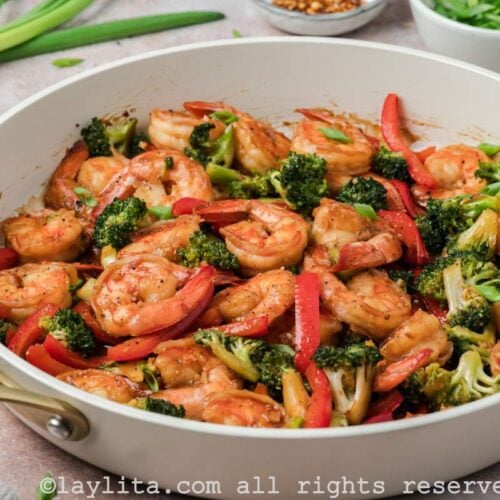 Side view of a skillet of shrimp broccoli stir fry with green onions and ginger in the background