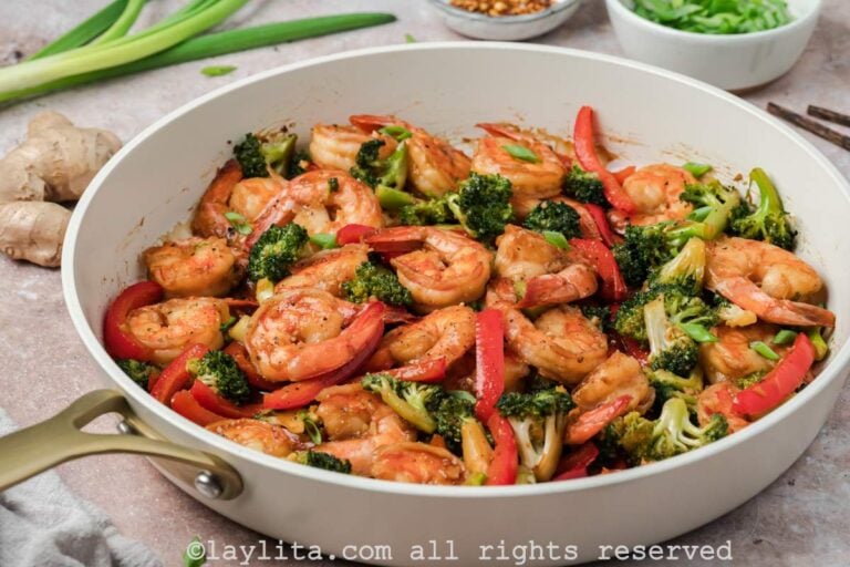 Side view of a skillet of shrimp broccoli stir fry with green onions and ginger in the background