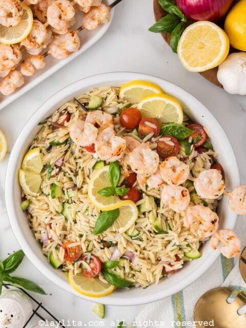 Overhead view of a bowl of orzo pasta salad topped with grilled shrimp, with more shrimp and vegetables in the background