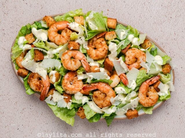 Overhead view of a platter with chopped romaine lettuce, croutons, cooked shrimp, parmesan and Caesar dressing