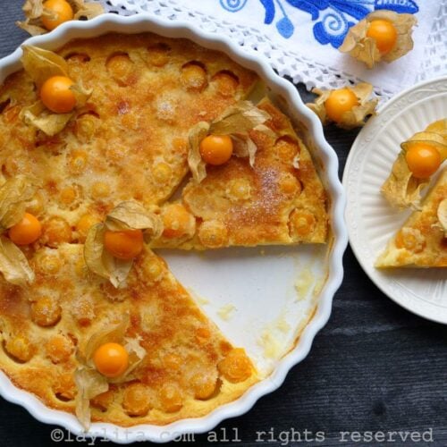 Overhead shot of uvilla clafoutis in a baking dish next to a slice of it on a plate with a blue floral napkin in the background