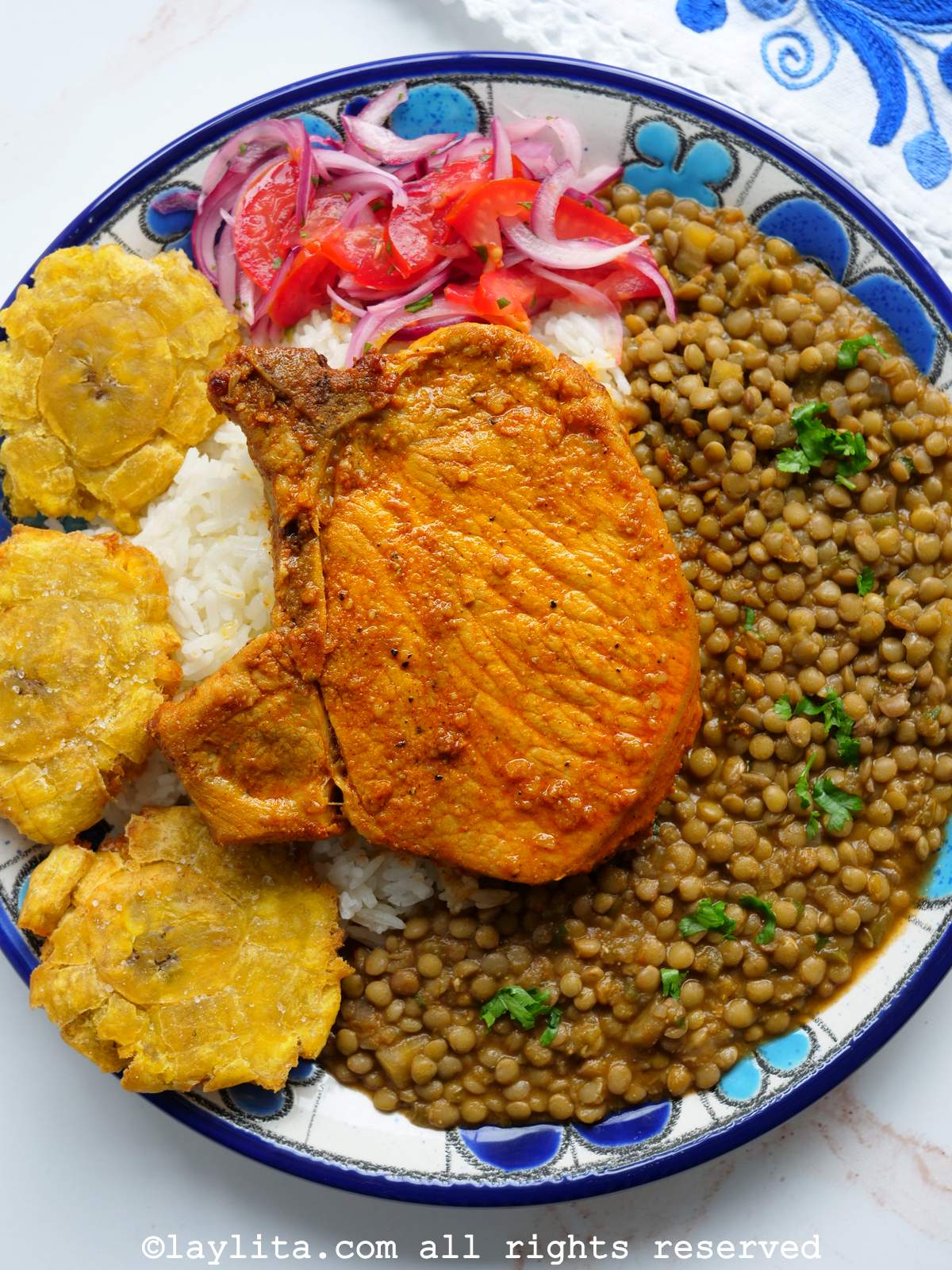 Overhead shot of an achiote pork chop served with rice and lentils on a blue and white plate on a white background.