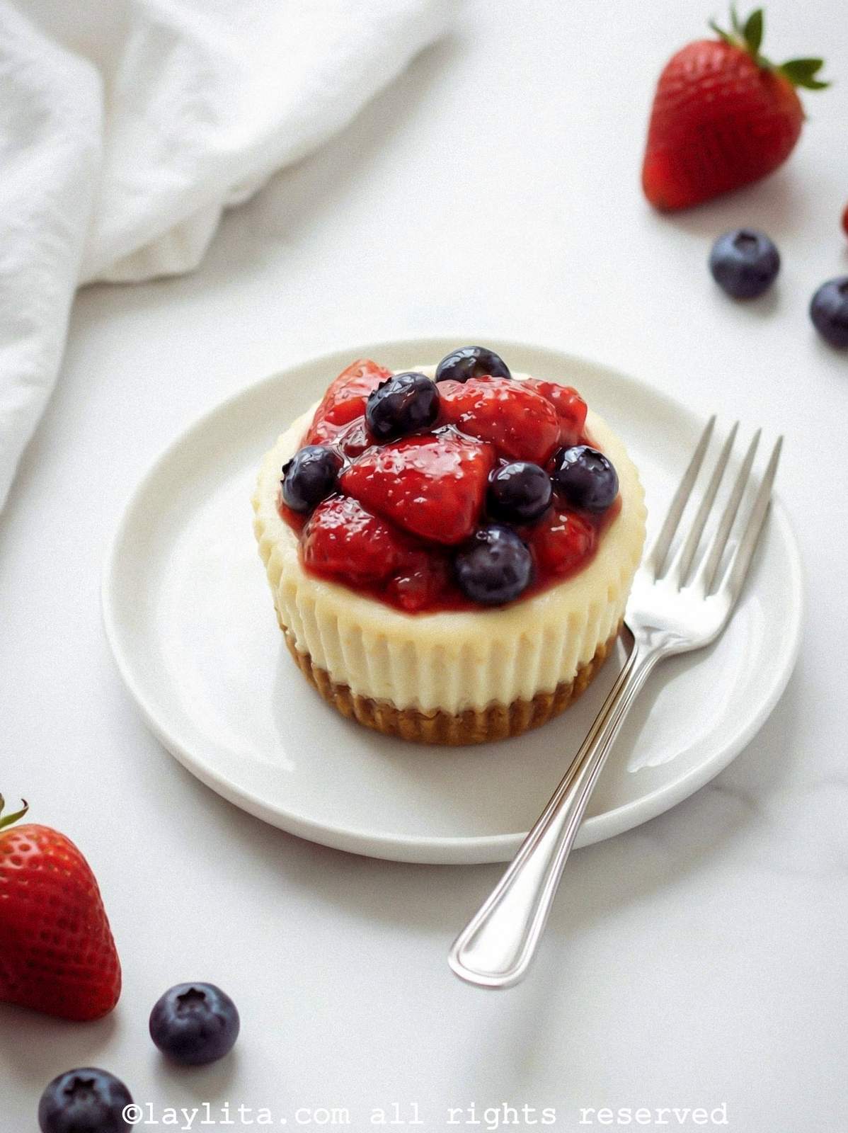 A mini cheesecake topped with berry sauce on a white plate with a fork next to it and berries on the side