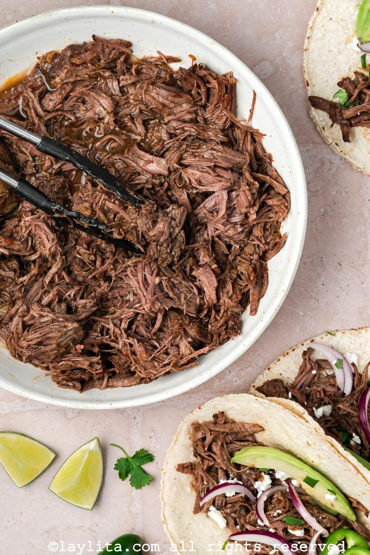 Overhead shot of three barbacoa tacos leaning on each other, next to a bowl of barbacoa beef.