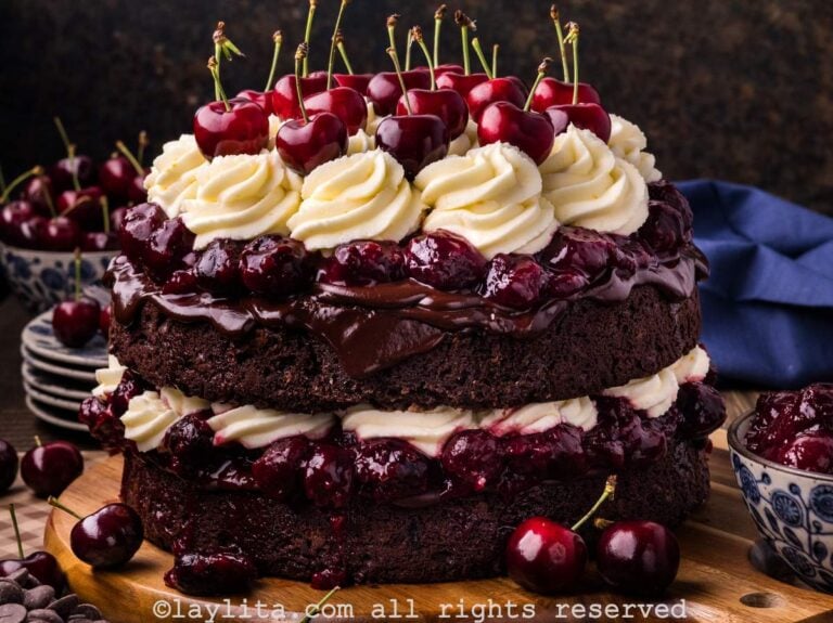 A layered Black Forest cake topped with whipped mascarpone frosting and cherries on a wooden platter with more cherries on the side