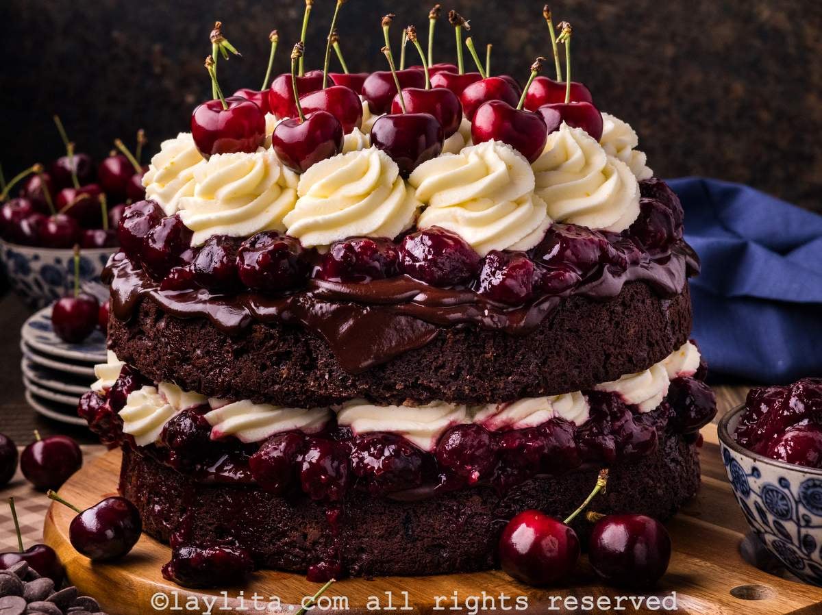 A layered Black Forest cake topped with whipped mascarpone frosting and cherries on a wooden platter with more cherries on the side
