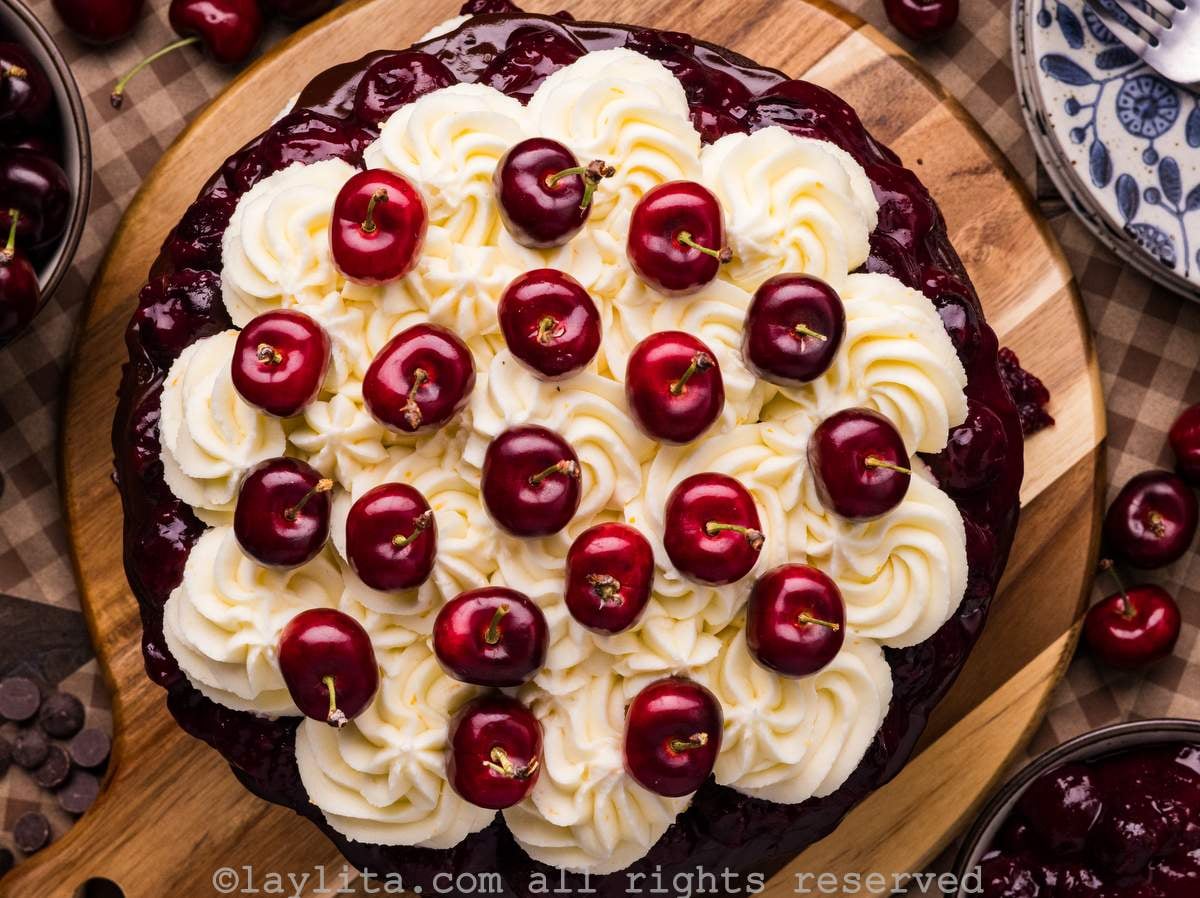 Overhead view of a Black Forest cake on a wooden platter showing the top part decorated with whipped cream and fresh cherries
