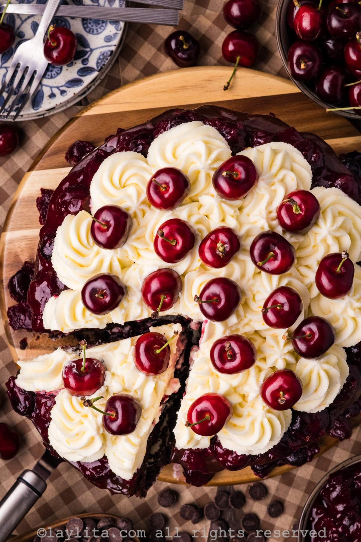 Overhead view of a Black Forest cake with a slice being removed on a wooden platter, showing the top part of the cake decorated with whipped cream and fresh cherries