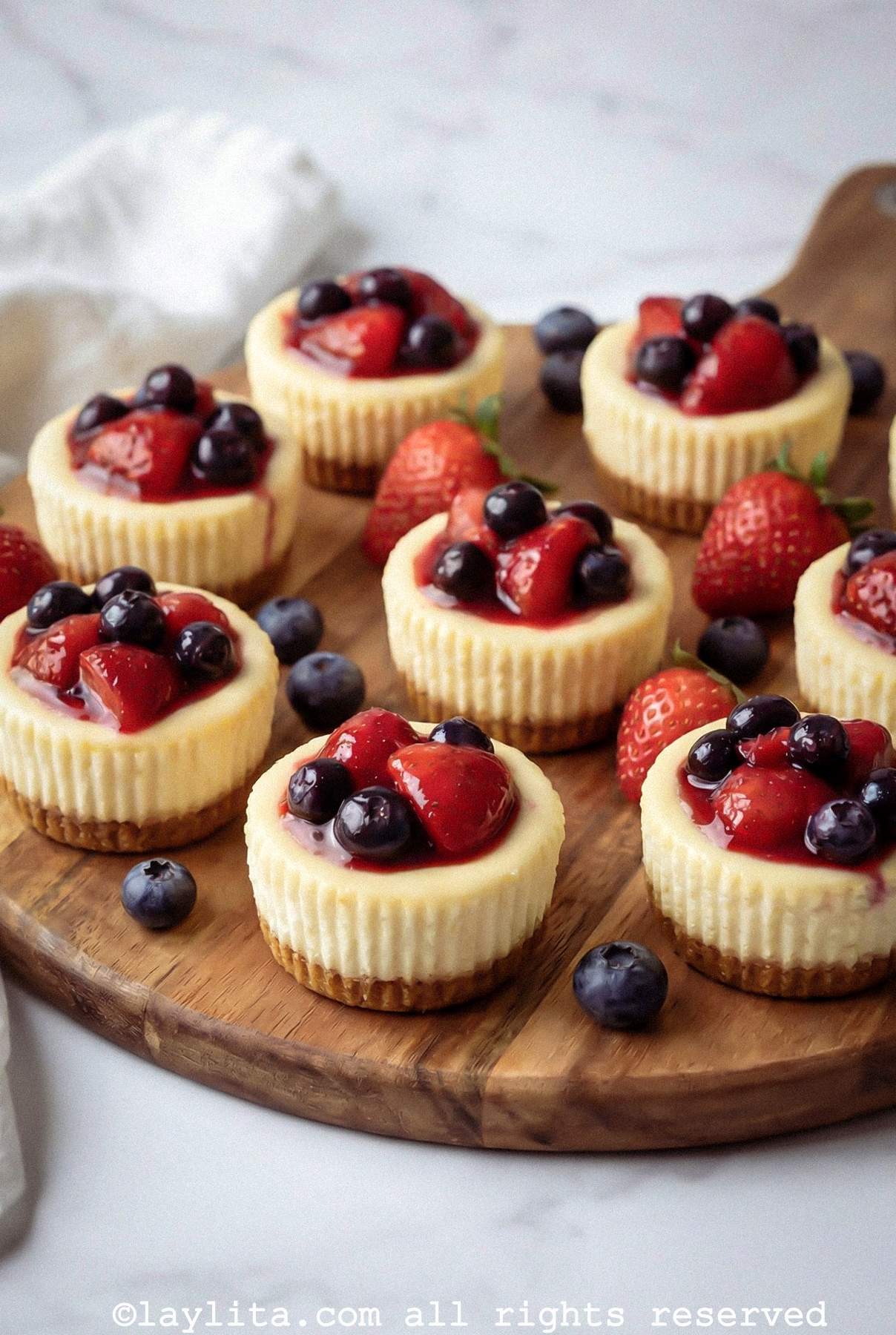 Mini cheesecakes on a wooden serving platter with fruit scattered around the plate.