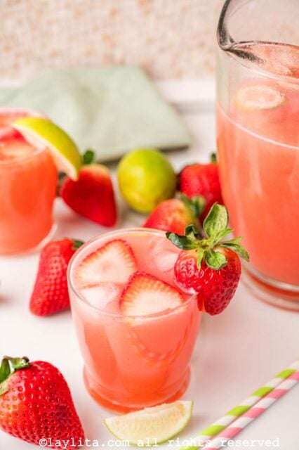 Glass of agua fresca with fresh strawberries on top, next to the pitcher of agua fresca.