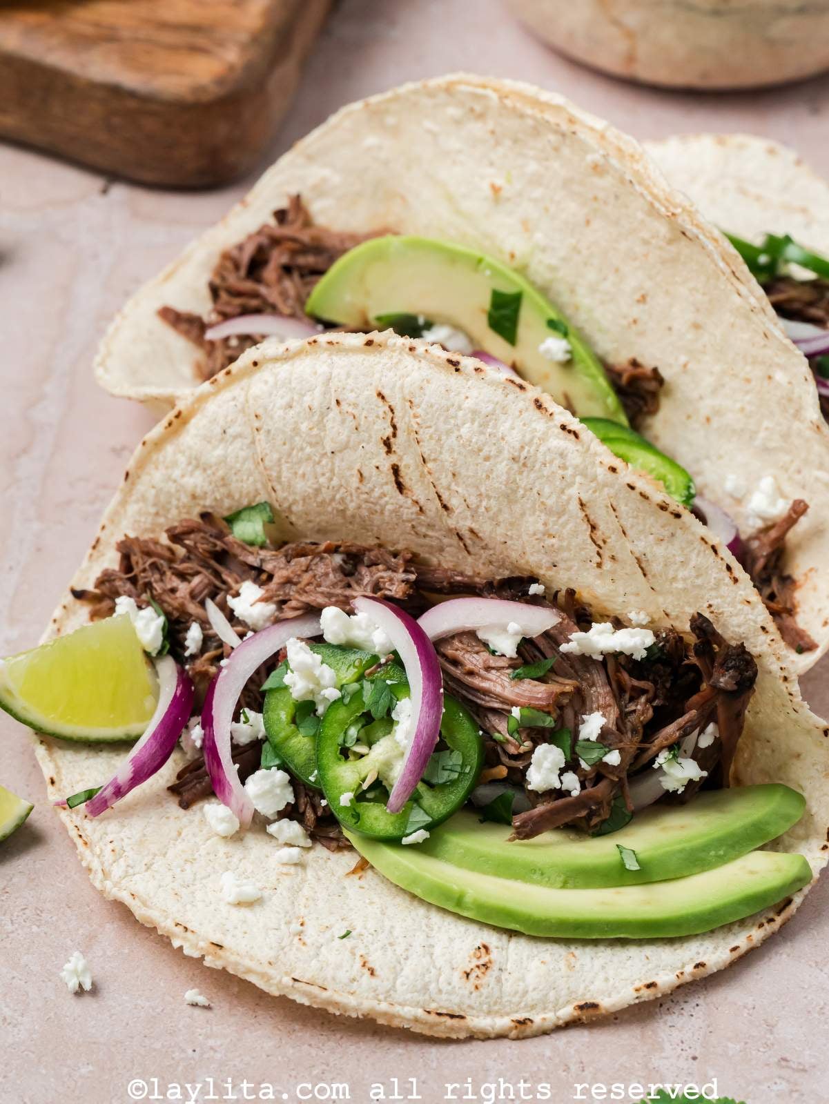 Beef barbacoa tacos on a plate with avocado, jalapeno, and red onions on top