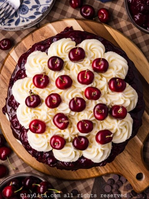 Overhead view of a Black Forest cake on a wooden platter showing the top part decorated with whipped cream and fresh cherries