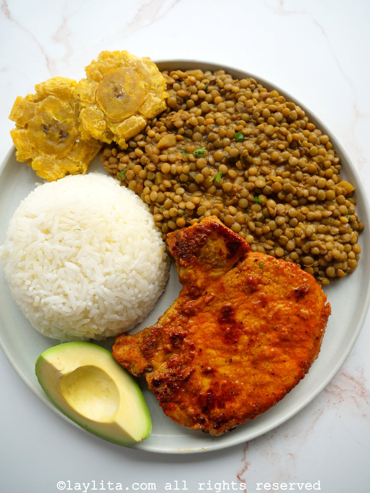 Achiote pork chop served on a white plate with lentils, rice, plantains, and avocado.