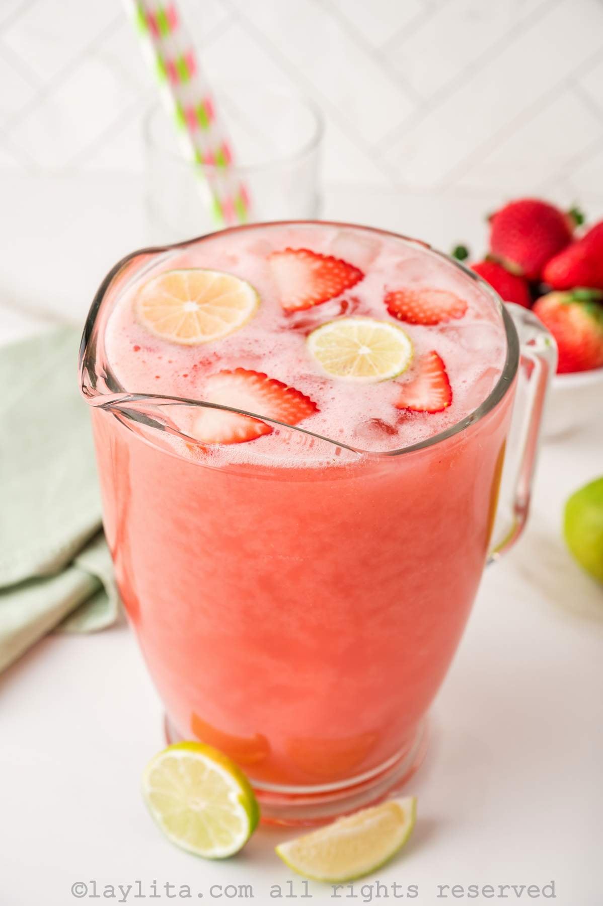 A pitcher of agua fresca with strawberries and limes in it, with more strawberries in the background